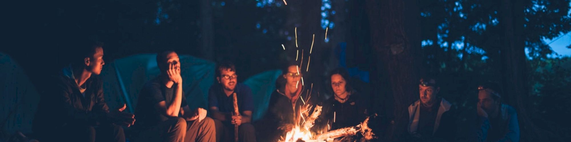 A group of people sitting around a campfire in a forest at night, with sparks flying up in the air.
