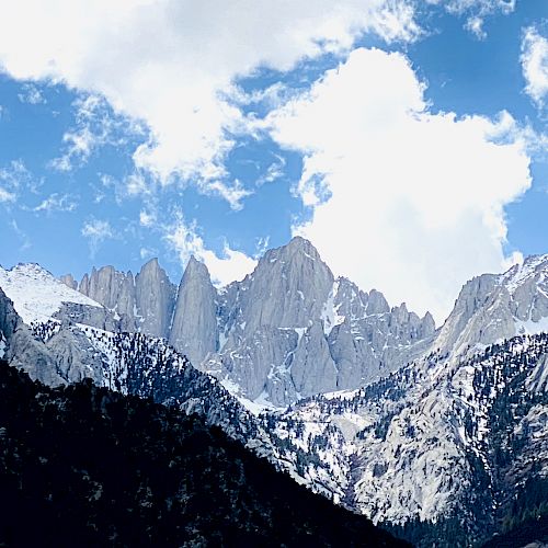 Snow-covered mountain peaks reach towards a partly cloudy, blue sky, framed by dark, rugged hills.