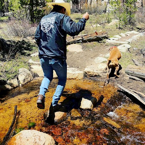 A person wearing a cowboy hat is jumping over a creek of clear water with rocks, followed by a dog in a wooded area.