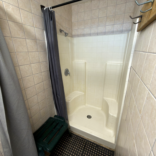 Small tiled shower stall with a white acrylic tub/shower, gray curtain, and a dark green bench on the tiled floor.
