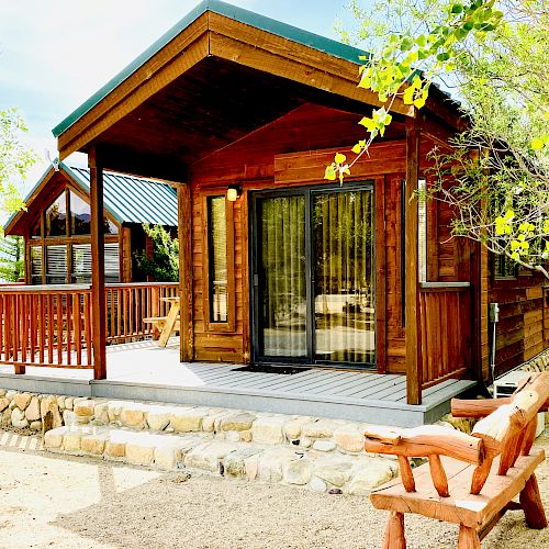 A cabin with a wooden porch, large windows, and a stone foundation, surrounded by trees and a wooden bench on the ground.