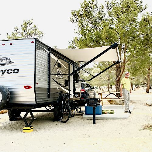 A person stands beside a silver Jayco RV parked at a campsite with an awning extended, surrounded by trees and camping gear, on a sunny day.