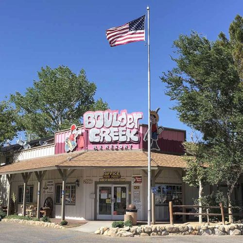 A small storefront with a wooden deck, an American flag atop, and a sign that reads &ldquo;BUILDERS CREEK&rdquo; in a rustic, western-style setting.