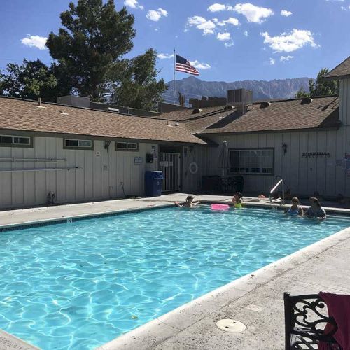 A sunny motel-style outdoor pool with light-blue water, lounge chairs, an American flag, and a few people relaxing by the pool.
