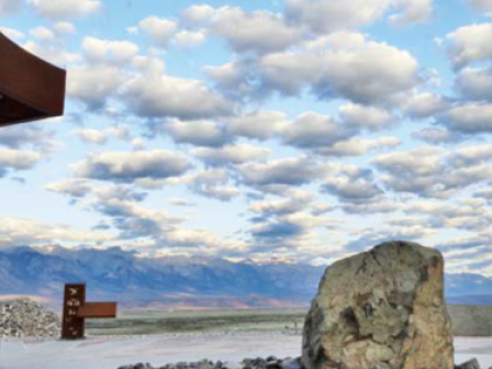 A scenic landscape with a large rock in the foreground, mountains in the distance, and a partly cloudy sky. There is also a metal structure on the left.