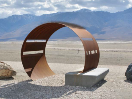 A large circular sculpture made of rusted metal stands in a barren desert landscape with mountains in the background.