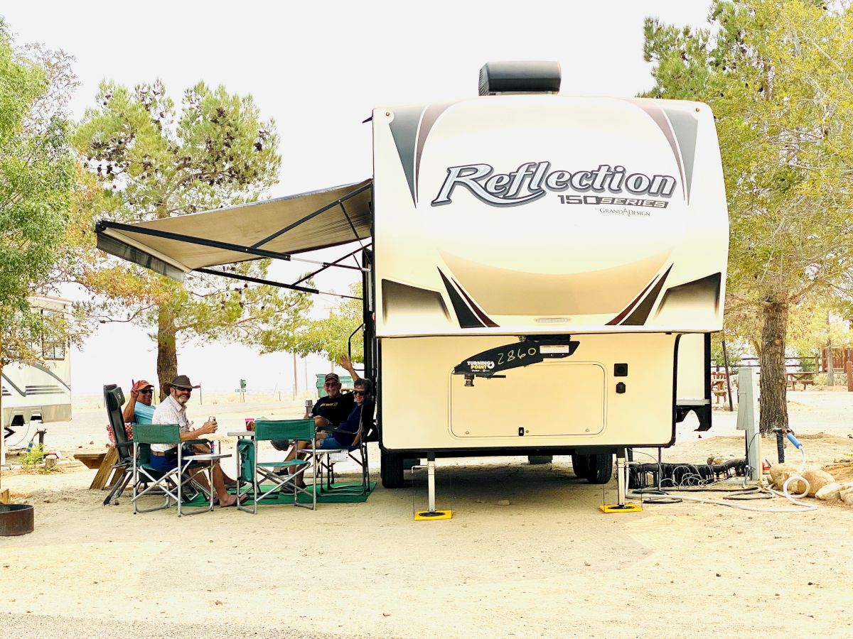 A large RV with an open awning, people seated under the shade, and trees in a desert-like campsite.