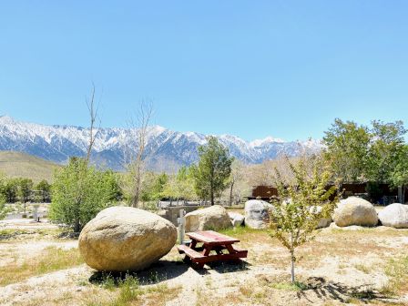 A sunny outdoor scene with large boulders, a picnic table, green trees, and distant snow-capped mountains under a clear blue sky.