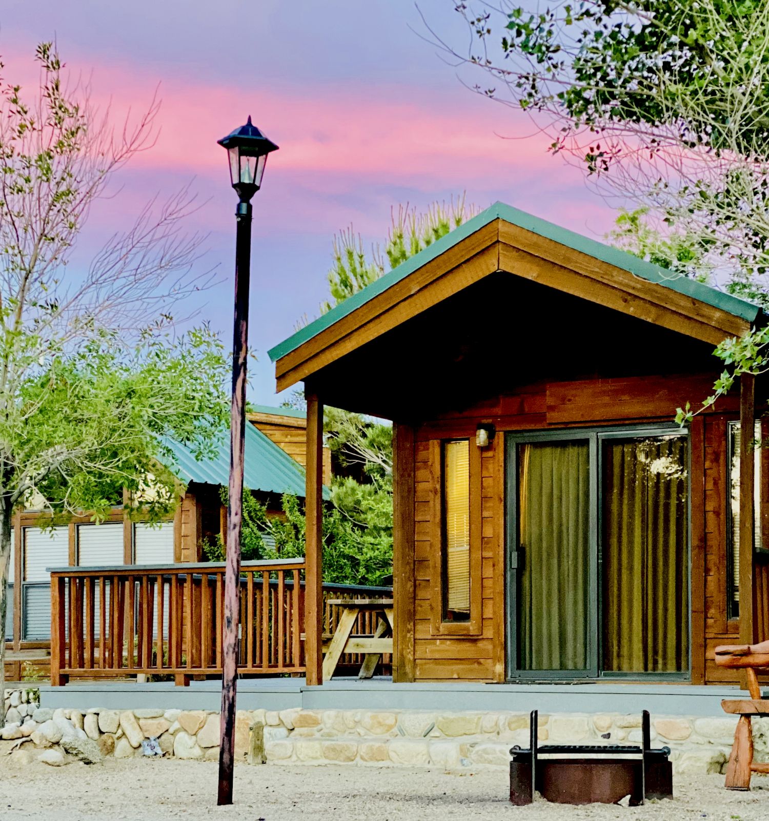 A cozy wooden cabin with a porch, outdoor seating, a stone path, and a lamp post against a pink sunset and leafy trees.