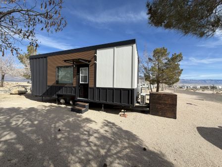 A modern tiny home trailer on a sandy lot near trees, with a small porch, stairs, and a clear blue sky.