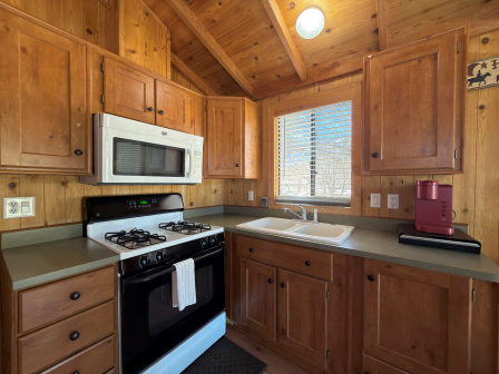 A cozy wooden kitchen with warm cabinetry, a white microwave, gas stove, double sink by a window, and a red coffee machine on the counter.