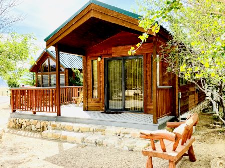 A cozy wooden cabin with a stone foundation, front porch, sliding glass doors, and a wooden bench on a sunny, tree-filled yard.