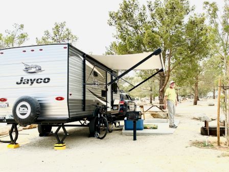 A Jayco travel trailer set up with an awning, outdoor chairs, and a man standing near a power setup in a sunny desert campground.