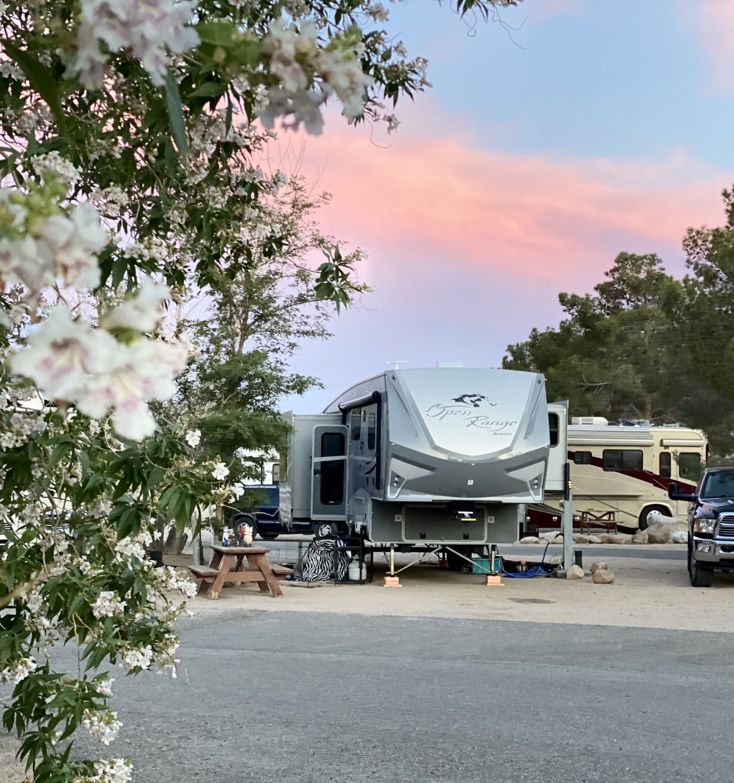 A campground with a large travel trailer, another RV, and a pickup truck parked among trees by a pastel sunset sky.