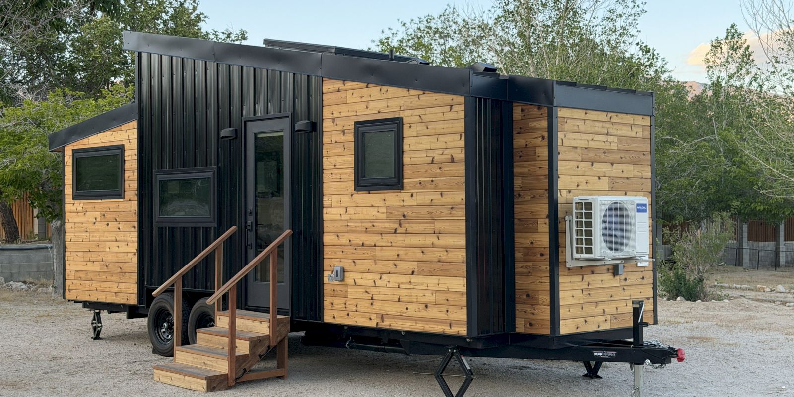 A modern tiny house on a trailer with wood siding, black steel accents, small steps to the door, and an exterior AC unit mounted on the side.