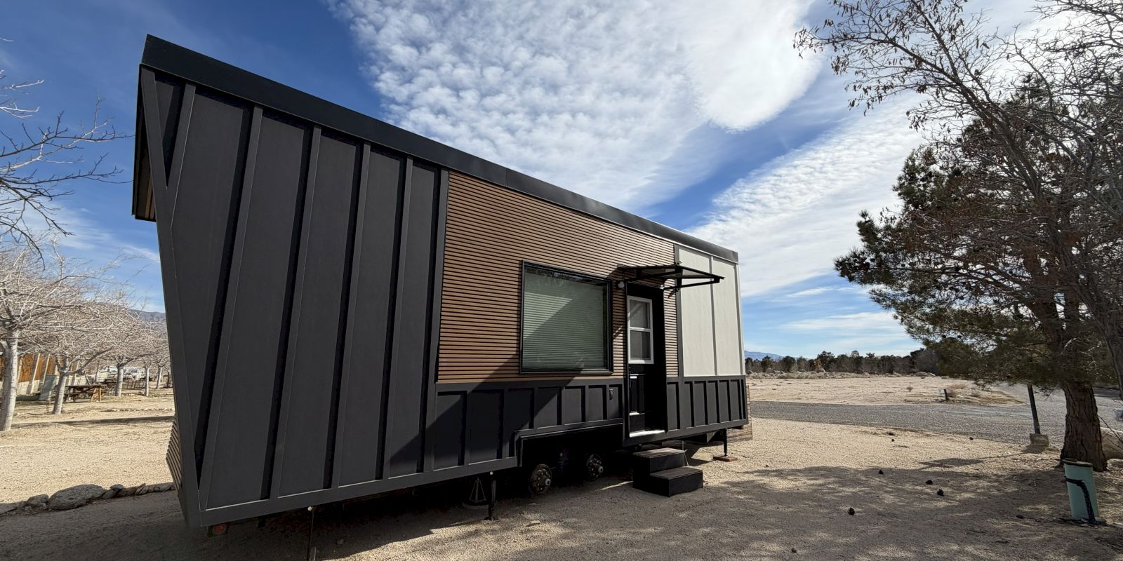 A small, black metal tiny house on wheels sits on a sandy lot with a clear sky, single tree, and a short ramp at the door.