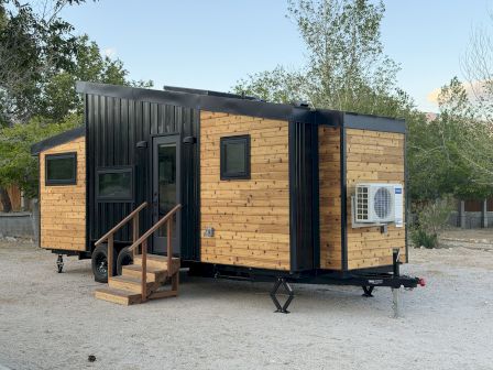 A modern tiny house on a trailer, with wood siding, black trim, small porch stairs, and an exterior air conditioner unit, parked on a gravel lot.