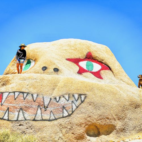 A giant desert rock sculpture with a teethy mouth, a red eye and a green iris; two people pose on its top under a bright blue sky.