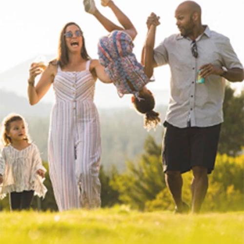 A family of four enjoys a sunny day outside with the parents swinging their daughter between them while the other child walks alongside.