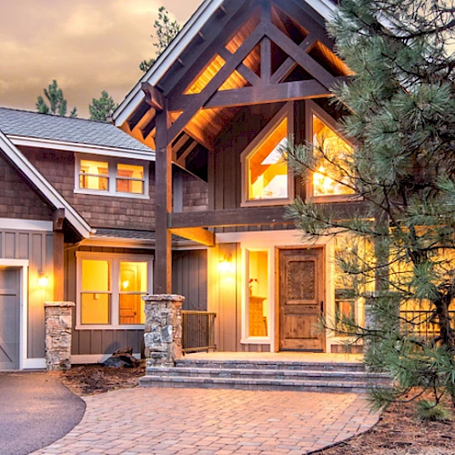 A spacious, rustic-styled house with glowing warm lights, multiple peaked roofs, a wooden exterior, and an attached double garage surrounded by trees.