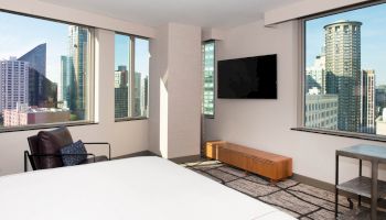The image shows a modern bedroom with large windows, city skyline view, a mounted TV, a chair, and minimalist furniture.