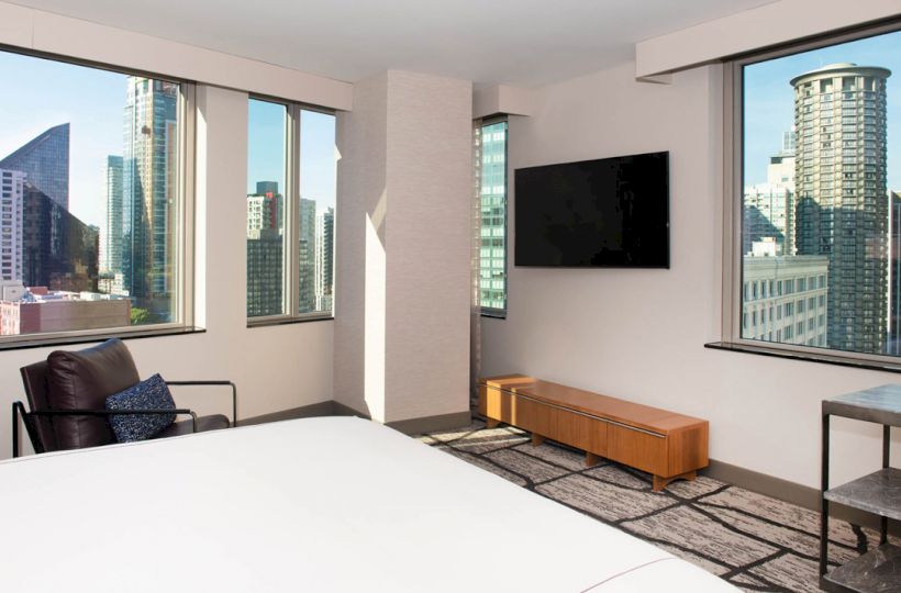 The image shows a modern bedroom with large windows, city skyline view, a mounted TV, a chair, and minimalist furniture.