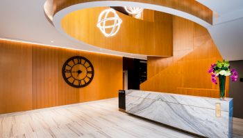The image shows a modern reception area with a curved wooden staircase, a clock on the wall, a marble counter, and a floral arrangement on the counter.
