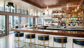 The image shows a modern bar with a long counter and high stools, a well-stocked liquor shelf, large windows, and an outdoor seating area.