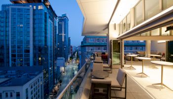 The image shows a modern cityscape at dusk, featuring a high balcony with tables and chairs, overlooking tall buildings and a street below.
