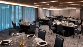 The image shows a banquet hall set up for an event, featuring round tables with black tablecloths, chairs, and place settings, under modern lighting.
