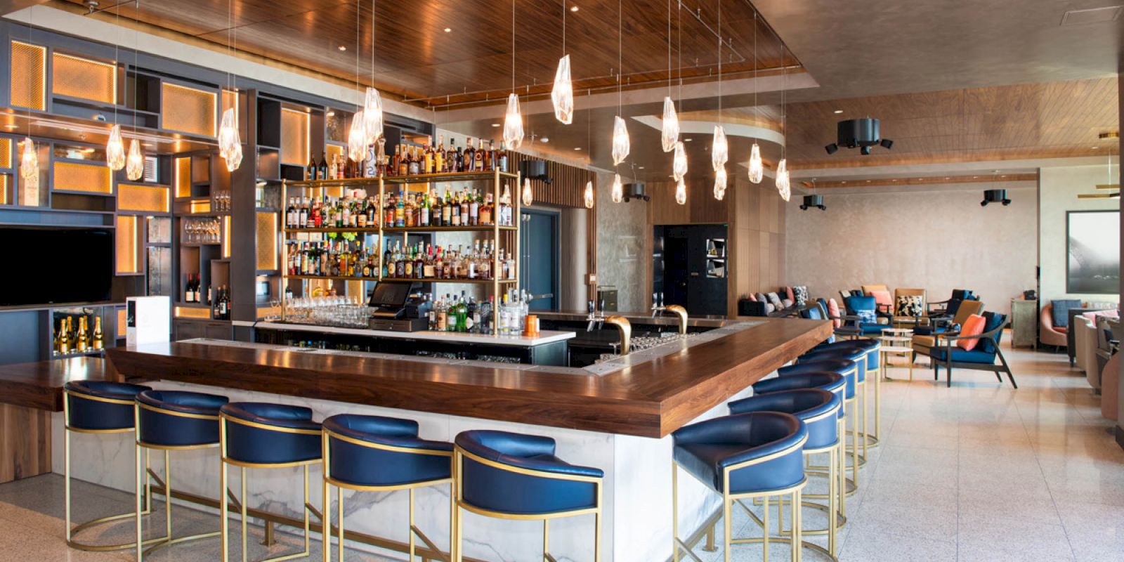 The image shows a modern bar with a large counter, numerous bottles behind it, elegant lighting, and blue bar stools.