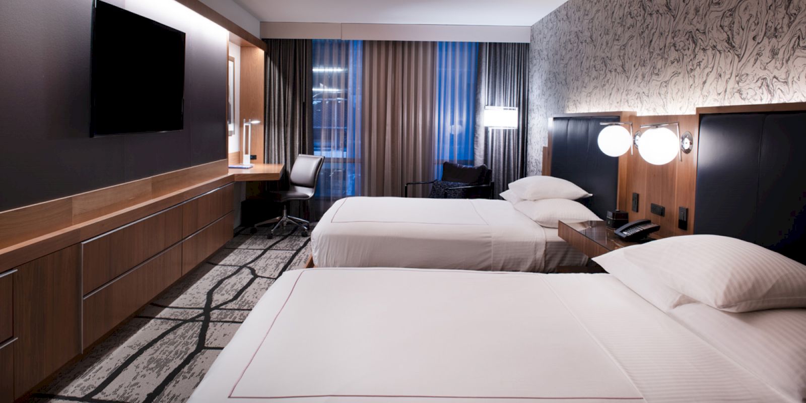 A modern hotel room with two beds, a flat-screen TV, a desk with a chair, and stylish lighting and decor.