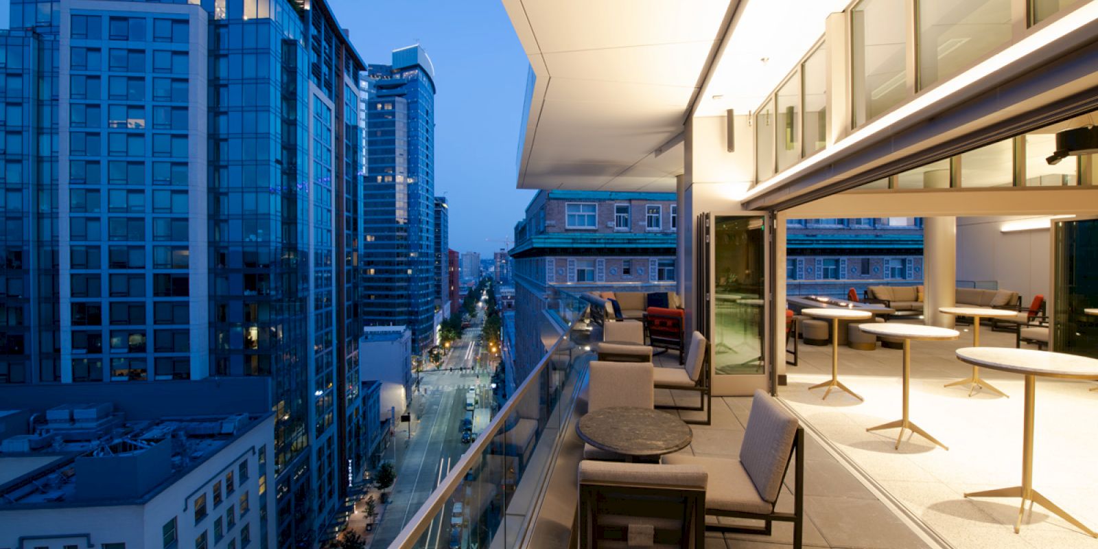 A night-time cityscape taken from a high balcony, showing modern buildings, a street lined with lights, and a furnished outdoor patio extending inside.