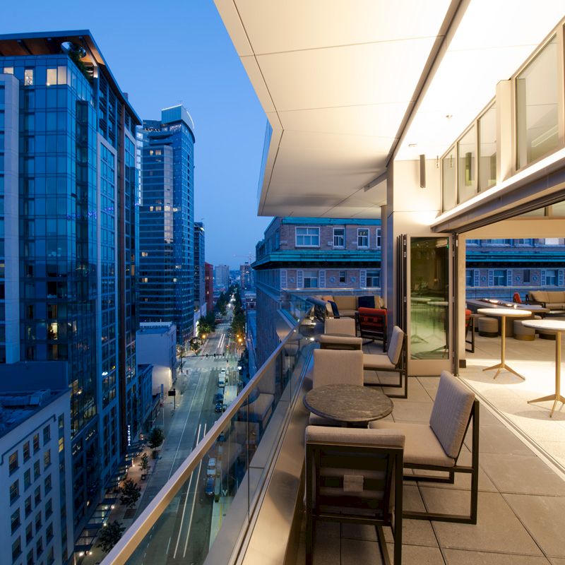 The image shows a modern balcony overlooking a city street with tall buildings, featuring outdoor seating and tables under evening lighting.