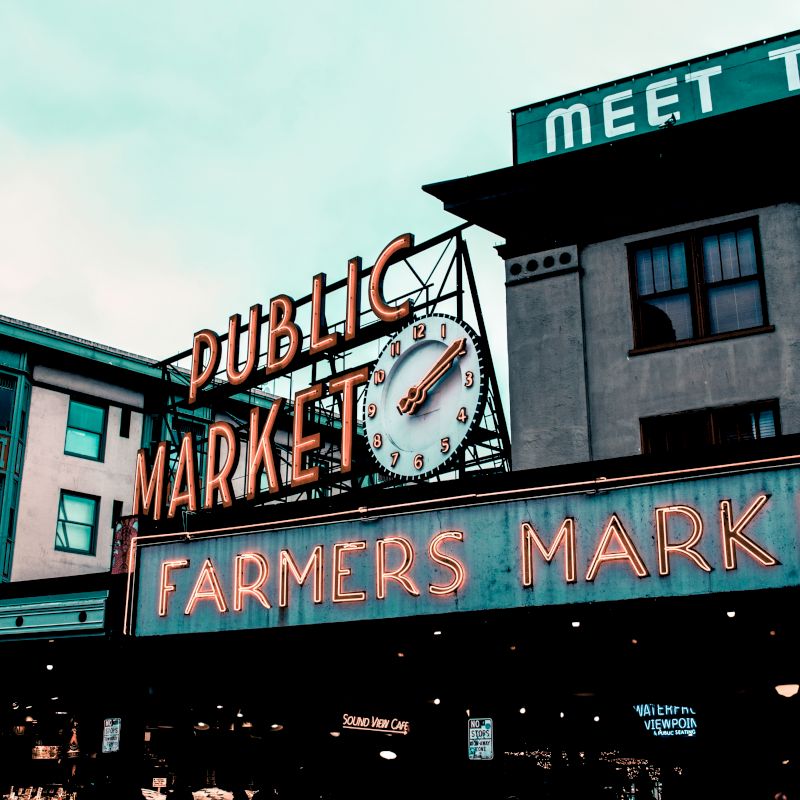 A public market sign with a clock and neon "FARMERS MARKET" text is displayed on a building, with surrounding buildings visible in the background.