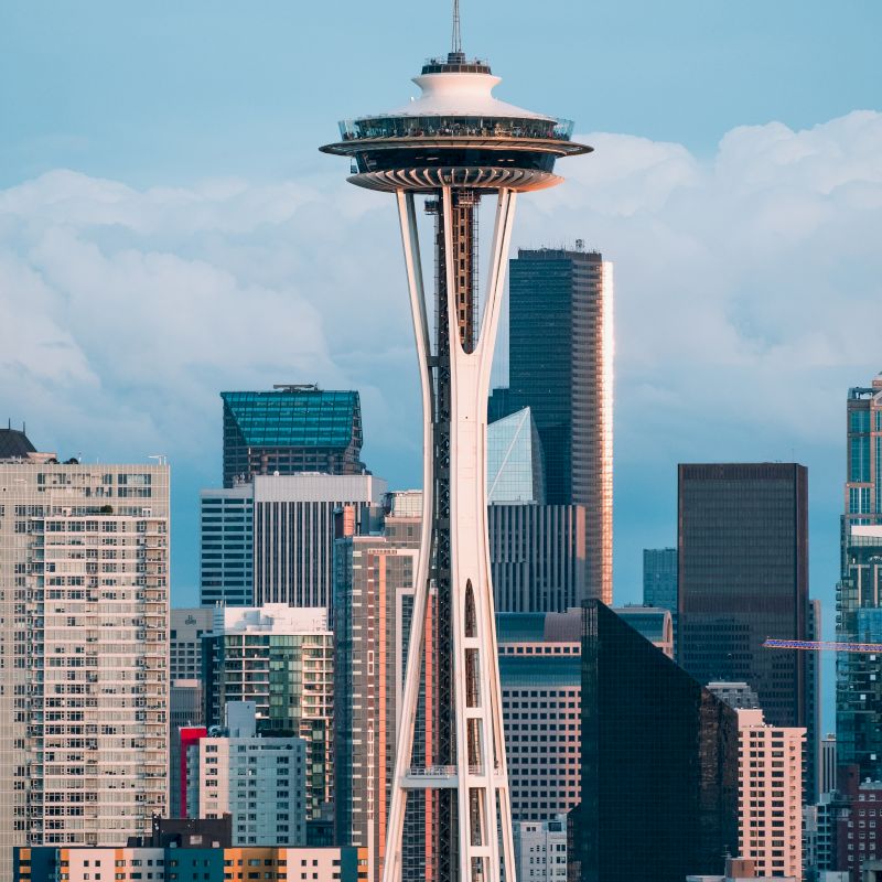 The image features the Seattle Space Needle towering over the city's skyline with various modern buildings and a partially cloudy sky in the background.