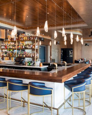 A modern bar with a wooden counter, blue chairs, hanging lights, and a well-stocked bar shelf. A lounge area is visible in the background.