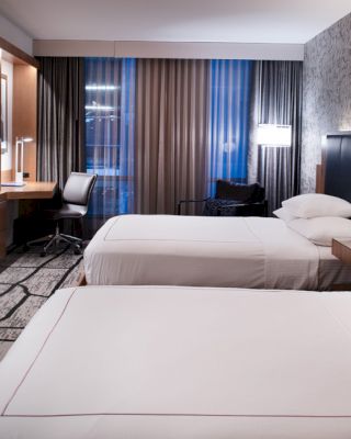 This image shows a modern hotel room with two neatly made beds, a desk and chair by the window, and a wall-mounted TV. The sentence ends here.