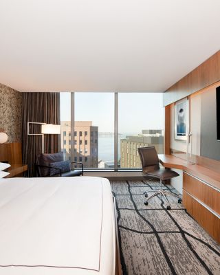 A modern hotel room features a large bed, a flat-screen TV, a desk, and a chair, with a window offering a cityscape view.