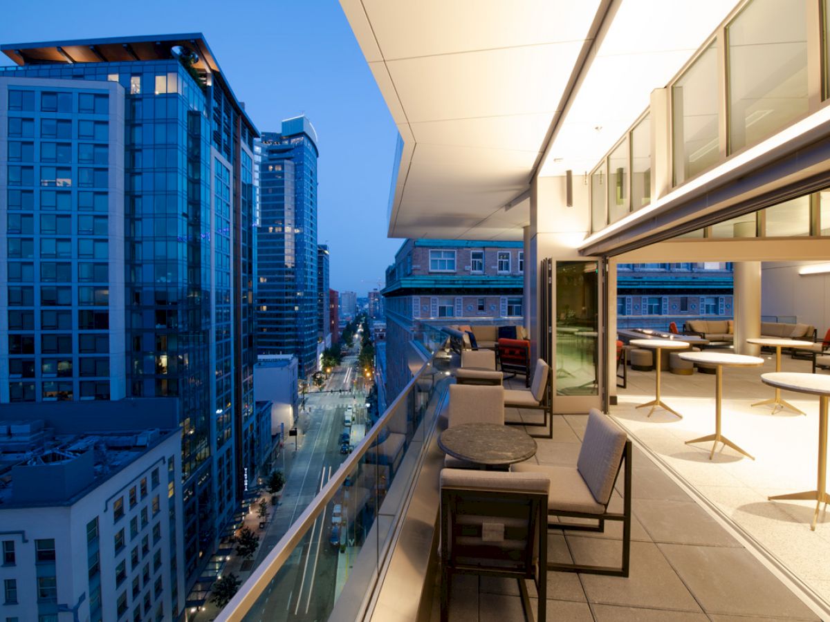 A modern cityscape at dusk featuring tall buildings with glass facades alongside a spacious balcony with chairs and tables.