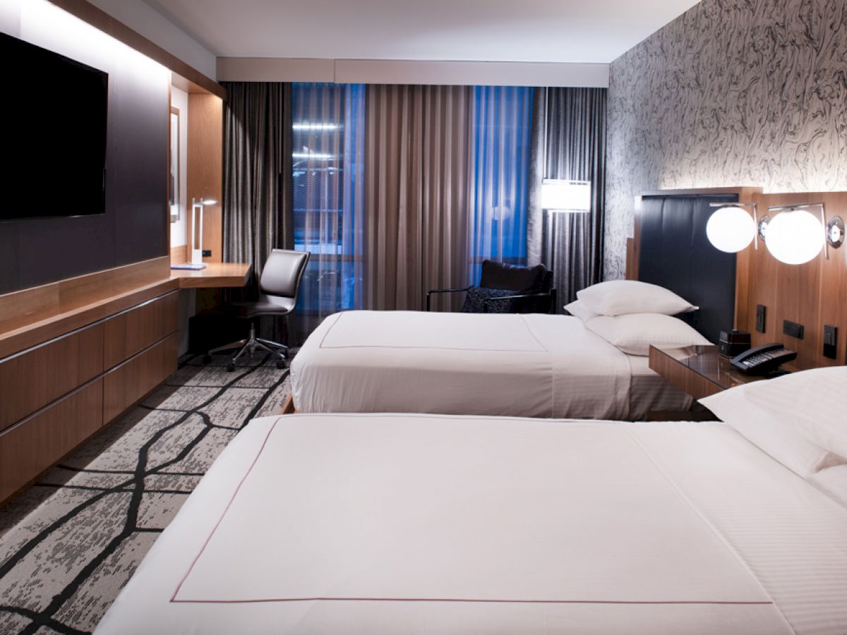 The image shows a modern hotel room with two neatly made beds, a desk and chair, a flat-screen TV, wall lamps, and floor-to-ceiling windows.
