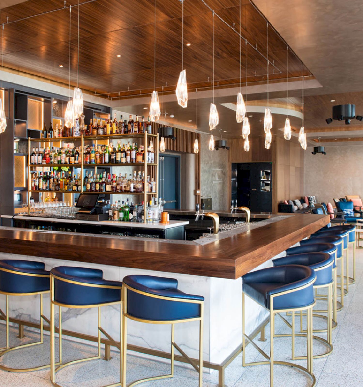 The image shows a modern, stylish bar with a well-stocked liquor shelf, pendant lights, blue bar stools, and a cozy seating area in the background.