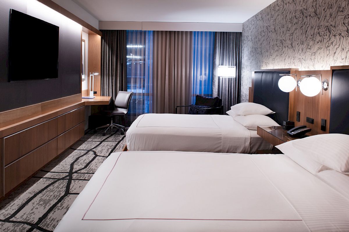 A modern hotel room with two beds, a flat-screen TV, a desk with a chair, bedside tables, and contemporary decor.