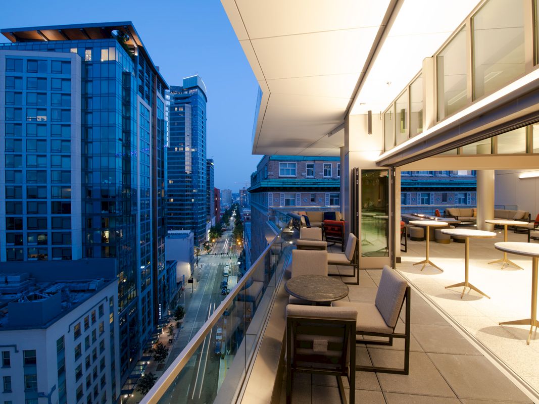 This image shows a modern cityscape at dusk with high-rise buildings. There is a balcony with outdoor furniture and tables on the right side of the image.