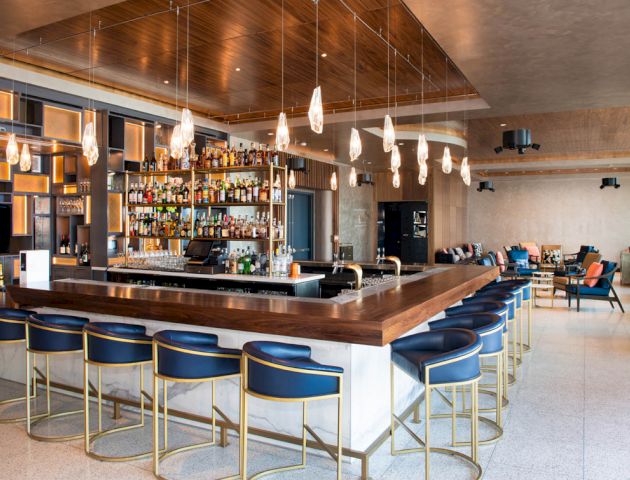 A modern bar with a wooden counter, blue bar stools, various bottles on shelves behind the bar, and hanging lights in a spacious, stylish setting.