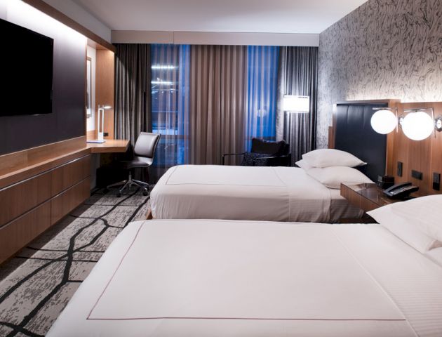 A modern hotel room with two neatly made beds, wall-mounted TV, desk with chair, and stylish décor with ample lighting.