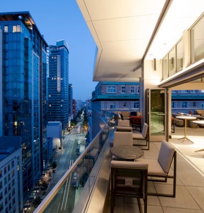 An upscale cityscape and terrace view at dusk, with modern high-rise buildings and stylish outdoor seating on the balcony.