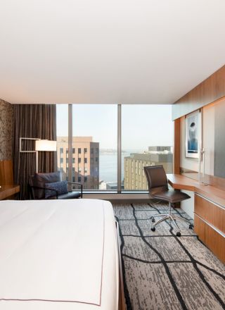 A modern hotel room features a large bed, wall-mounted TV, desk, and window offering a city view. The decor is contemporary with artistic wallpaper.