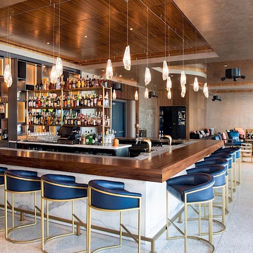 This image shows a modern bar with blue barstools, hanging lights, a fully stocked bar, and additional seating areas in the background.