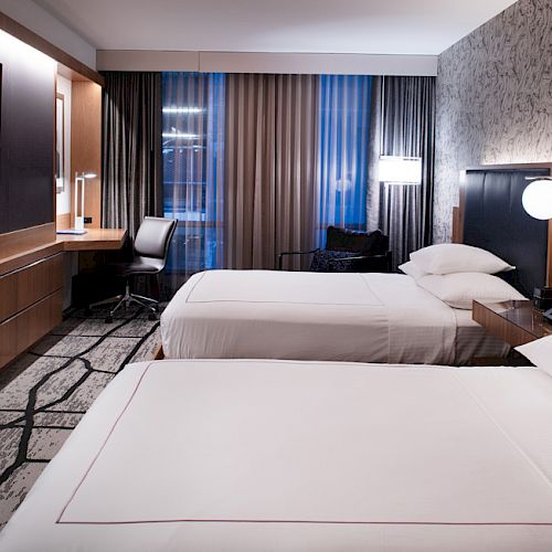 A modern hotel room with two beds, a desk, a chair, wall-mounted TV, and stylish lighting. Curtains cover the windows in the background.